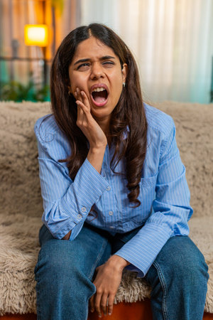 Indian young woman presses cheek in pain from sudden toothache while sitting on home sofa, expression strained and tense. Arabian girl shows clear dental discomfort and distress from oral health issueの写真素材