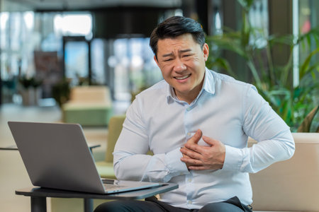 Stressed Chinese mature businessman using laptop and suffers from tension holding chest with heart...