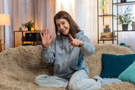 Portrait of smiling young woman joyfully exclaims Hey you and waves hand palm in hello hi gesture welcomes someone with hospitable expression expresses positive emotions. Girl sitting on sofa at homeの写真素材