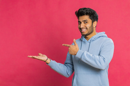 Indian bearded man showing thumbs up and pointing left empty place, advertising area for commercial text copy space for goods promotion advertisement. Guy isolated on red background. Lifestylesの写真素材