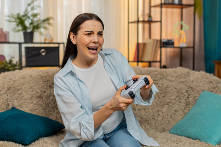Young excited woman at home sitting on sofa playing video game using joystick emotionally reacting...