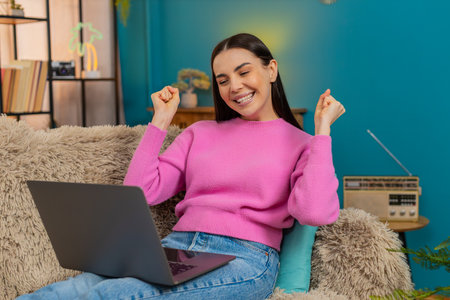 Young woman at home raising hands up with excitement and joy celebrating successfully completed laptop project. Caucasian girl on sofa happy, showing satisfaction, achievement and positive emotionsの写真素材