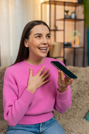 Young woman at home talking on smartphone using speaker mode, emotionally sharing story with gestures. Caucasian girl on sofa expressing thoughts with hands, full of energy, passion and connectionの写真素材