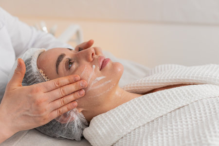 Caucasian woman girl lies relaxed as cosmetologist gently massages her cheeks with cleansing foam during skincare routine. Young woman enjoys professional beauty facial treatment in clinic. Horizontalの写真素材