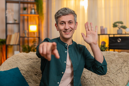 Young happy man at home pointing at camera with smile during video call greeting excitedly. Caucasian guy on sofa shows joy happy energy and enthusiasm, saying hello with expressive friendly attitudeの写真素材