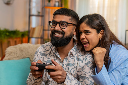 Indian couple man and woman sitting on home sofa with game controller and popcorn, laughing and shouting while playing console game. Young family enjoying fun emotional moment with excitement indoors.の写真素材