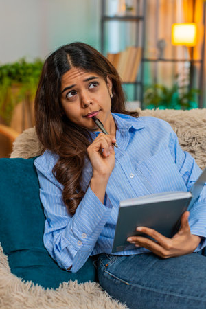 Indian young woman writes in diary while on home sofa with light thoughtful smile and relaxed posture. Arabian girl reflects on day with peaceful emotion, personal time and soft inner calm indoors.の写真素材