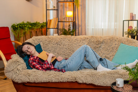 Young woman lies on home sofa holding book, slowly falling asleep with relaxed body and tired expression. Girl enjoys quiet end of day resting peacefully after reading with calm mind emotional comfortの写真素材