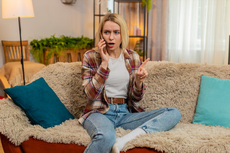 Angry adult woman rejects annoying call on smartphone while sitting on sofa, waving hand with refusal. Girl reacts with frustration to spam or scam caller, expressing disagreement with body language.の写真素材