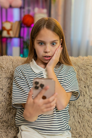 Young child girl sits on sofa reads surprising message on smartphone, reacts with wide open eyes curious expression. School kid looks stunned by unexpected content holding device tightly in both handsの写真素材