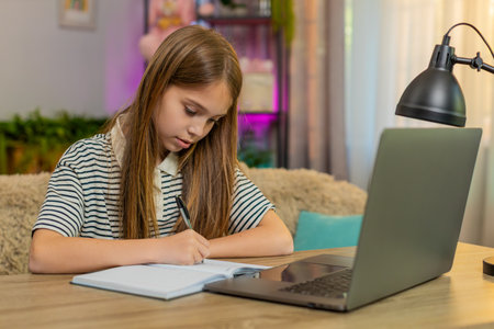 Child girl sitting at table on home couch attentively watches online lesson takes notes in notebook. School kid focuses on math homework, carefully writing numbers with deep concentration and effort.の写真素材