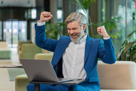 Caucasian bearded businessman in formal clothing working on laptop shocked by sudden victory, lottery goal achievement, good career news. Excited guy clenching fists sitting in modern hotel lobby.の写真素材