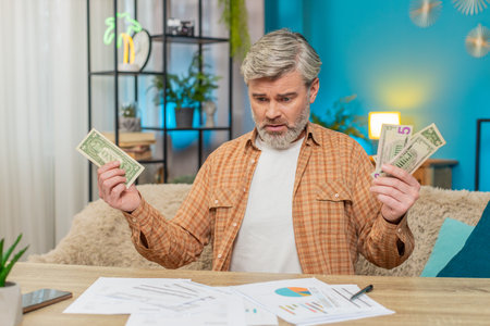 Mature man sitting at home table, surrounded by documents bills, holding a small amount of money...