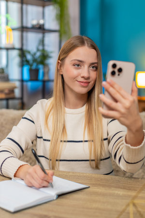 Caucasian woman in a serious business video call at home at table, holding smartphone writing notes. Girl appears focused determined calmly handling negotiations seeking solutions feeling professionalの写真素材