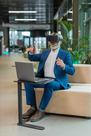 Mature businessman programmer software developer using headset app making a presentation. Watching virtual reality 3D 360 video. Bearded gray-haired guy in suit in VR goggles working at office lobby.の写真素材