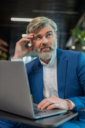 Bored sleepy mature businessman working on laptop computer, leaning on hand in modern office lobby....
