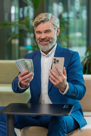 Successful happy rich mature businessman holding smartphone and counting dollar bill money fan sitting in office lobby. Happy Caucasian bearded guy winning online lottery game. Calculating budget.の写真素材