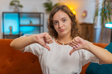 Dislike. Portrait of upset woman showing thumbs down sign gesture, expressing discontent, disapproval dissatisfied bad work at home apartment indoors. Displeased girl in living room on sofa. Lifestyleの写真素材