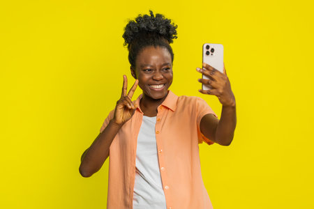 African American woman holds smartphone, smiling and posing for a selfie with cheerful energy. Black girl isolated on yellow background radiates confidence and happiness, capturing joyful momentsの写真素材