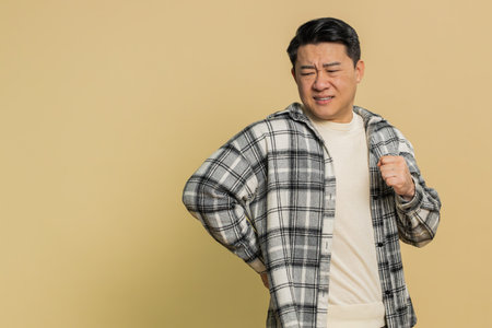 Asian middle-aged man suffering backache, massaging sore place lower spine, frowning from unbearable spasm torn muscles and tendons, kidney stones pinched nerve. Mature Chinese guy on beige backgroundの写真素材