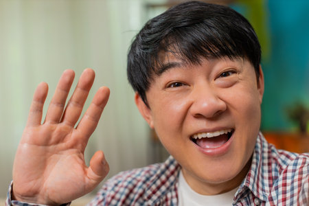 Asian man at home laughs, gesturing during POV video call, enjoying lively talk with friend. Chinese...