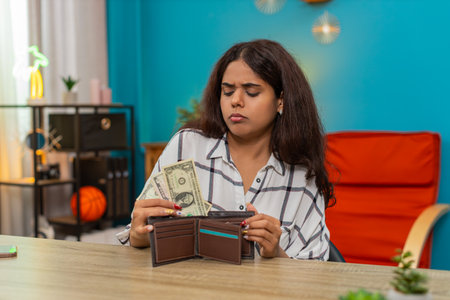 Indian woman at home opens wallet seeing almost no dollars cash money feeling deeply disappointed worried. Arabian sad girl at table holds few bills showing stress frustration worry budget anxietyの写真素材