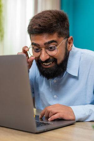 Indian man at home reads email reply on laptop smiles nods satisfied project finished success...