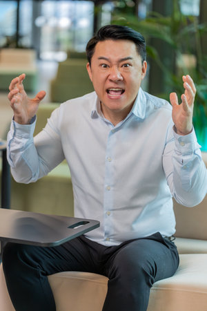 Angry mature boss in shirt arguing over video call looking at camera in modern workplace in office. Unsatisfied businessman manager communicate with subordinates about the company problems.の写真素材