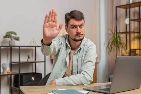 Young man freelancer with serious expression doing stop gesture with hand at home office desk,...