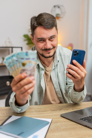 Planning family budget. Smiling man counting money Ukrainian hryvnias UAH cash use smartphone calculate domestic bills. Guy satisfied of income and saves salary money at home office table workplace.の写真素材