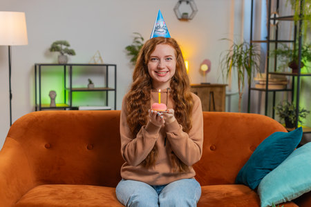 Happy woman wears festive birthday hat holding cupcake makes wish joyful congratulating blowing...