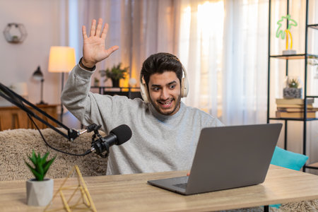 Young Indian man looking into laptop and webcam using headphones and studio microphone, recording vlog, conducting online stream and greeting subscribers while sitting at table in living room at home.の写真素材