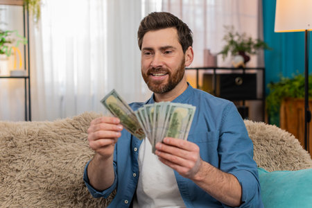 Young man at home holding a stack of dollar cash banknotes, counting money with excitement....