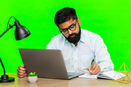 Man sits at desk writing ideas in notebook while working on laptop, showing focus, planning,...