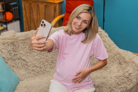 Pregnant middle-aged woman at home make video call selfie on smartphone to show belly smiling...
