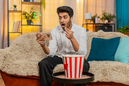Man on home sofa eating popcorn while watching a video on smartphone, fully engaged in content. Guy...