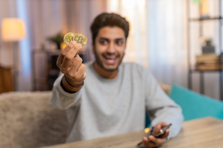 Young Indian man with bitcoin checks cryptocurrency exchange charts on smartphone app calculates...