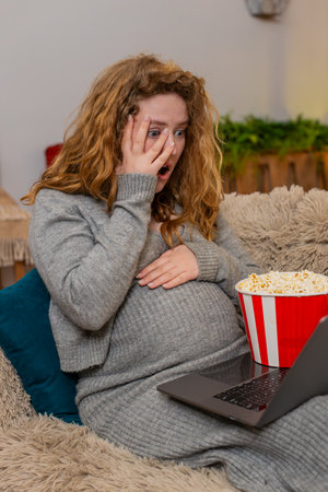 Pregnant young woman sits on home sofa, eating popcorn while watching scary movie on laptop,...