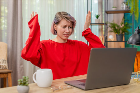 Young woman emotionally arguing with laptop screen sitting at home table, waving hand angrily...