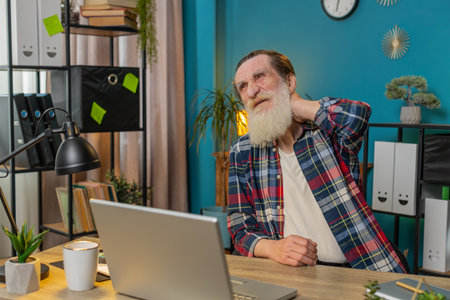 Tired Caucasian senior man suffers from spine neck muscle pain at home office workplace working on laptop netbook. Overworking. Bearded freelancer grandfather attempts to relieve pain by massaging.の写真素材