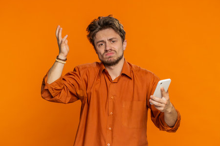 Upset sad Caucasian man use smartphone typing browsing, play game, loses, surprised by sudden lottery results bad fortune loss fail unlucky news. Handsome bearded guy isolated on orange backgroundの写真素材