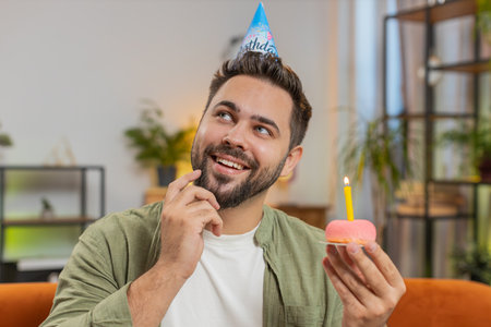 Happy man wears festive birthday hat holding cupcake makes wish joyful congratulating blowing burning candle on cake. Young brunette guy celebrating birthday party at home room apartment on couch.の写真素材