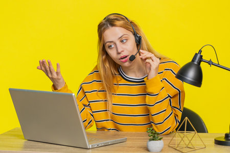 Young woman wearing headset, freelance worker, call center or support service operator helpline, having talk with client or colleague communication support. Girl sitting at table on yellow background.の写真素材