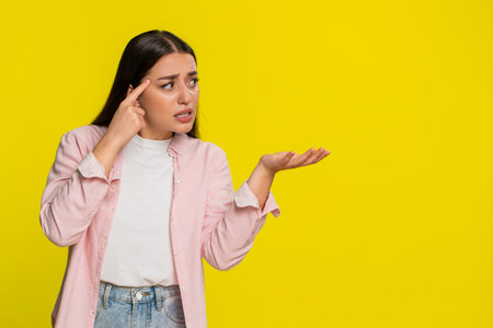 Young woman tapping finger on temple with sarcastic face, clearly expressing disbelief and criticism of an idea. Girl on yellow background shows reaction to nonsense and lack of smart thinkingの写真素材
