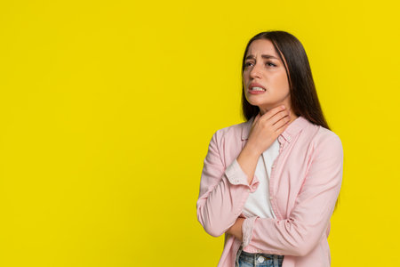 Young woman pressing hand to throat and squinting in pain, clearly showing discomfort while...