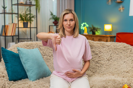 Pregnant middle-aged woman at home shows thumbs down after losing dislike gesture feeling...
