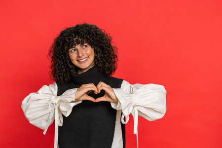 Woman in love. Smiling attractive Caucasian young woman with curly hair makes heart gesture demonstrates love sign expresses good positive feelings and sympathy. Girl isolated on red backgroundの写真素材