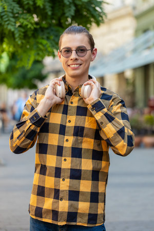 Joyful relaxed overjoyed young man choosing, listening favorite energetic disco rock n roll music dancing outdoors. Adult guy taking off wireless headphones looking at camera smiling on city street.の写真素材