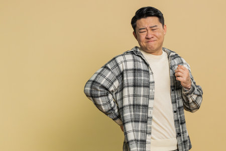 Asian middle-aged man suffering backache, massaging sore place lower spine, frowning from unbearable spasm torn muscles and tendons. Mature Chinese guy on beige backgroundの写真素材