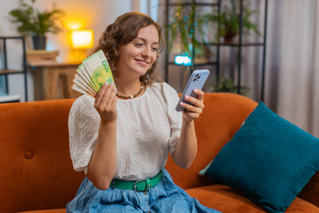Successful woman holding smartphone and waving money fan sitting on sofa couch at home. Happy girl winning online lottery game planning vacation calculating budget concept. Lifestylesの写真素材
